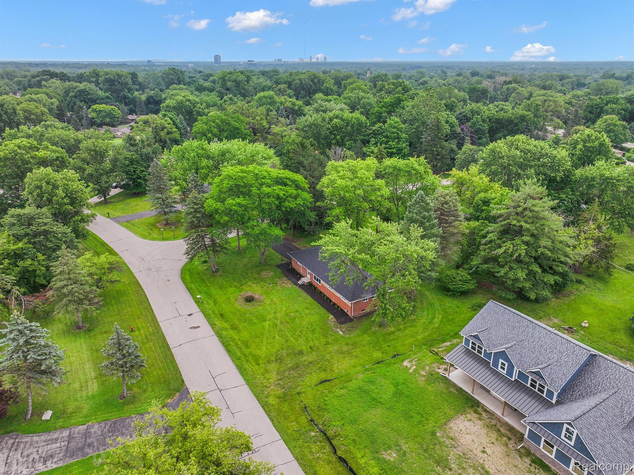 Aerial view of a brick home on a tree-lined residential street, with a broad lawn and distant skyline.