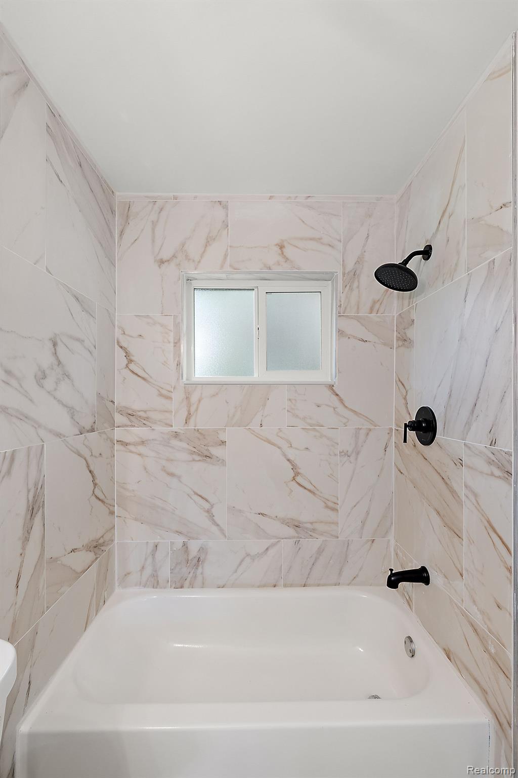 Bathroom tub beneath a frosted window, surrounded by veined tile with black fixtures.