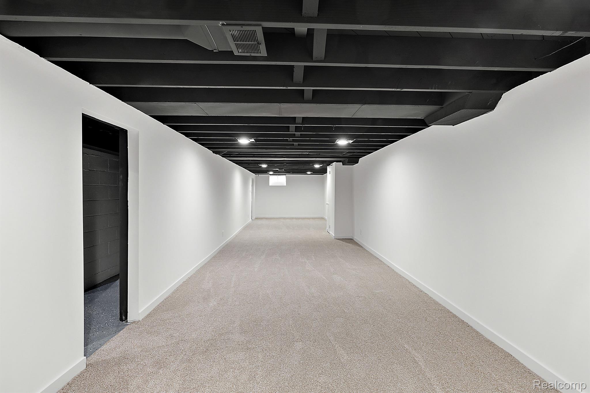 Vacant basement room with light carpet, white walls, and exposed black ceiling beams.
