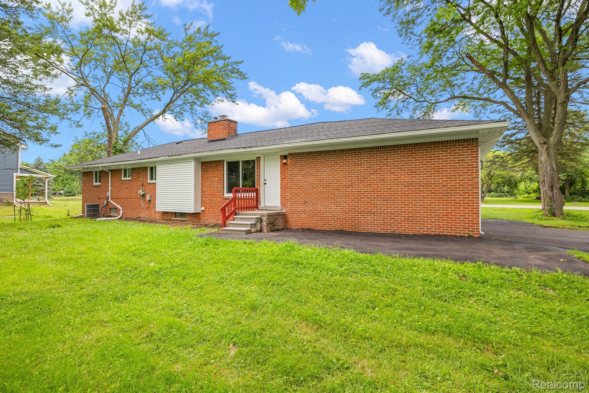 Red brick house with a white entry door and red steps, surrounded by a broad lawn and mature trees.
