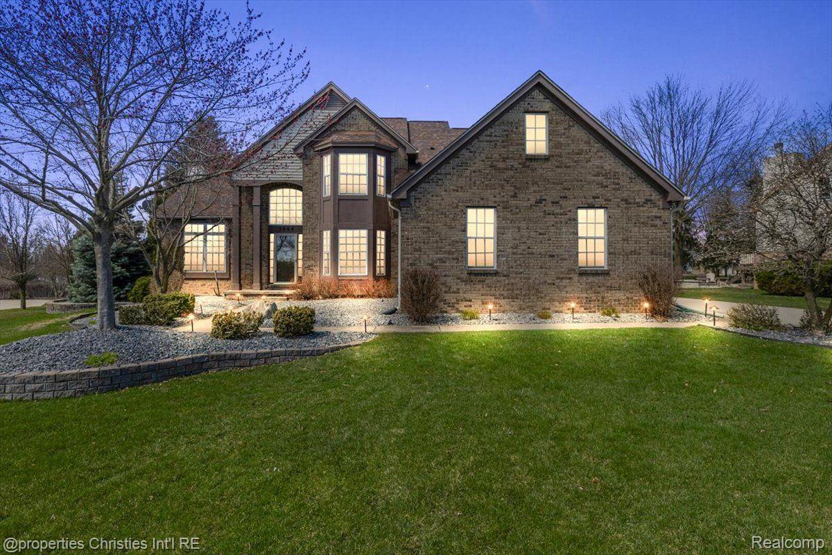 Brick house with steep gables, glowing windows, landscaped beds, and a broad lawn at dusk.