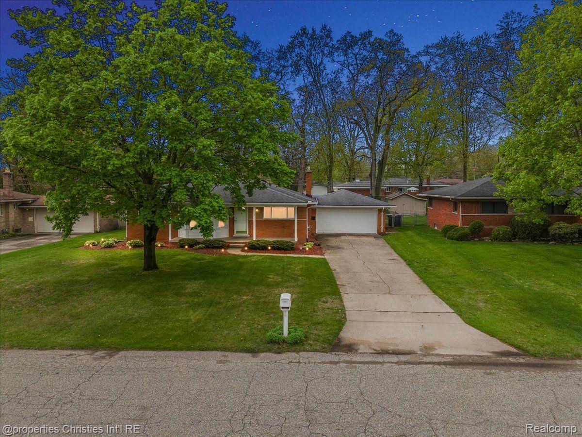 Single-story brick home with attached garage, broad driveway, and mature trees framing the front lawn.