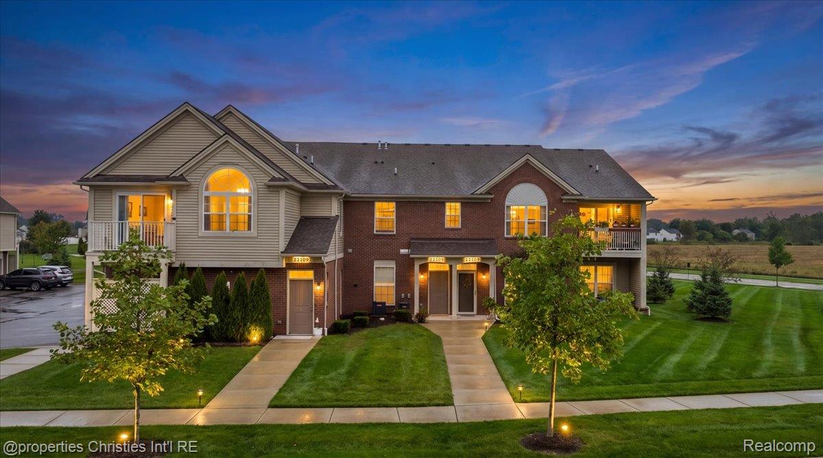Multi-unit residential building with brick and siding, lit windows, balconies, and landscaped lawns at dusk.