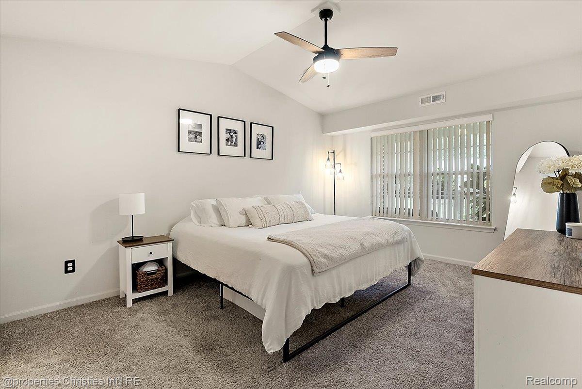 Staged bedroom with a white bed, gray carpet, ceiling fan, and large window with vertical blinds.