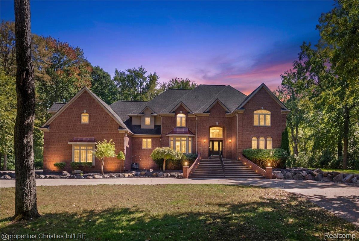 Large brick home with a broad front staircase, lit windows, and a colorful dusk sky.