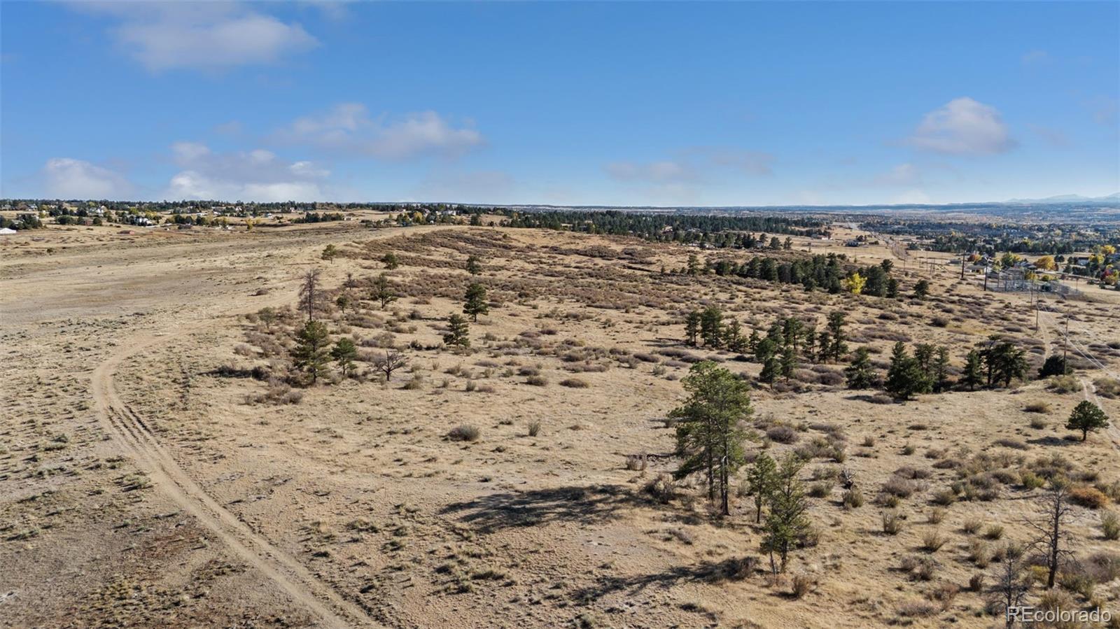 Aerial view of open land with scattered trees and a dirt track, with homes in the distance.