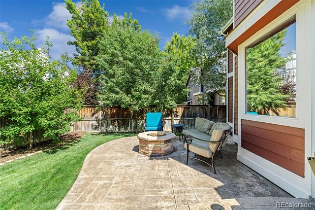 Backyard patio with a stone fire pit, cushioned chairs, and a fenced lawn shaded by mature trees.
