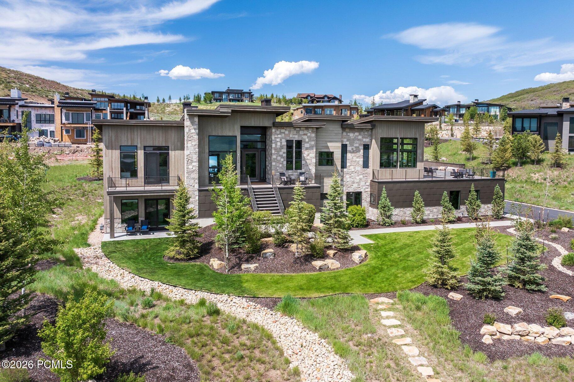 Modern stone-and-gray hillside home with broad terraces, curved lawn, and young evergreen trees.