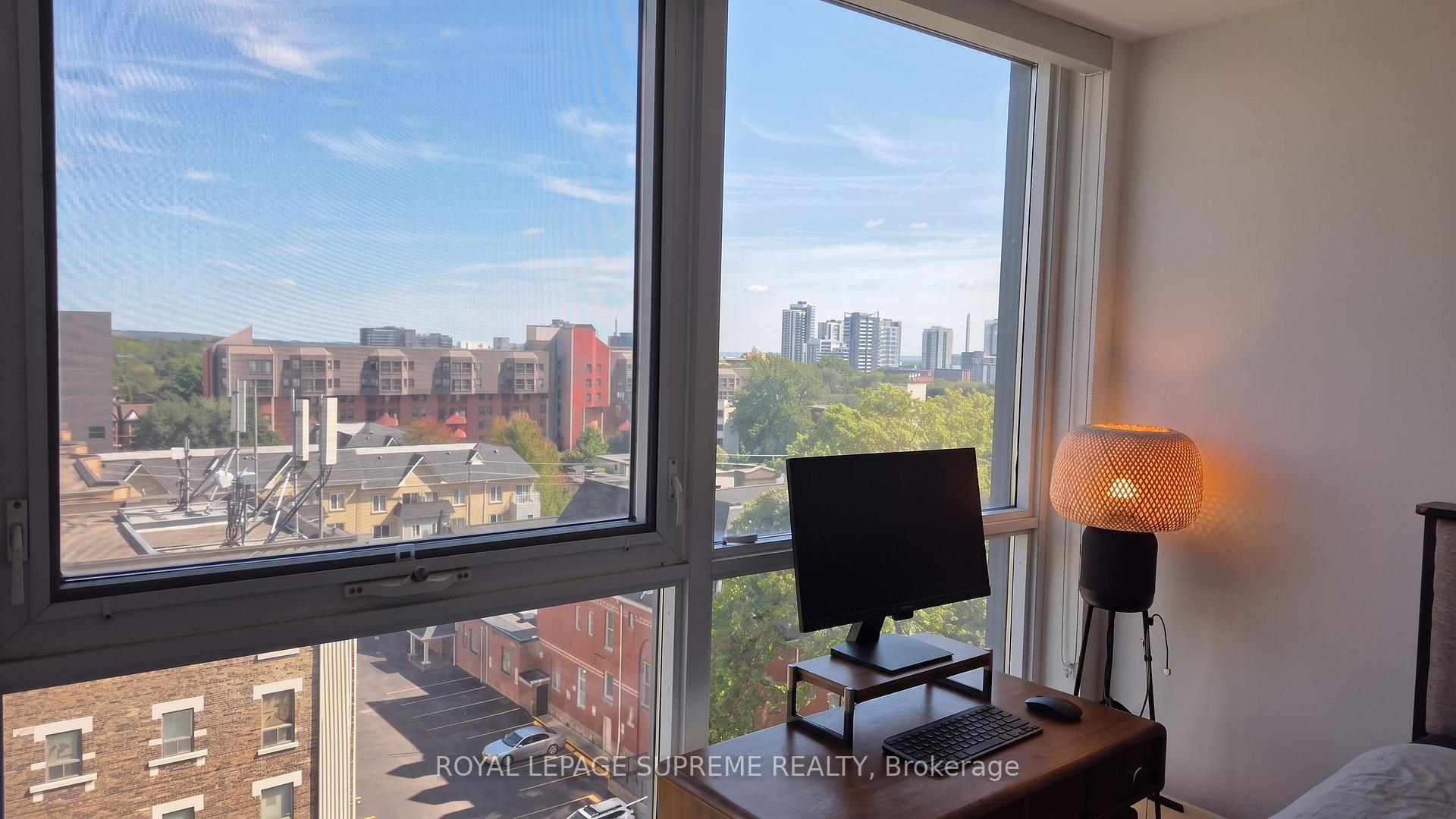 Condo bedroom with a desk beside large corner windows overlooking Toronto buildings and treetops.