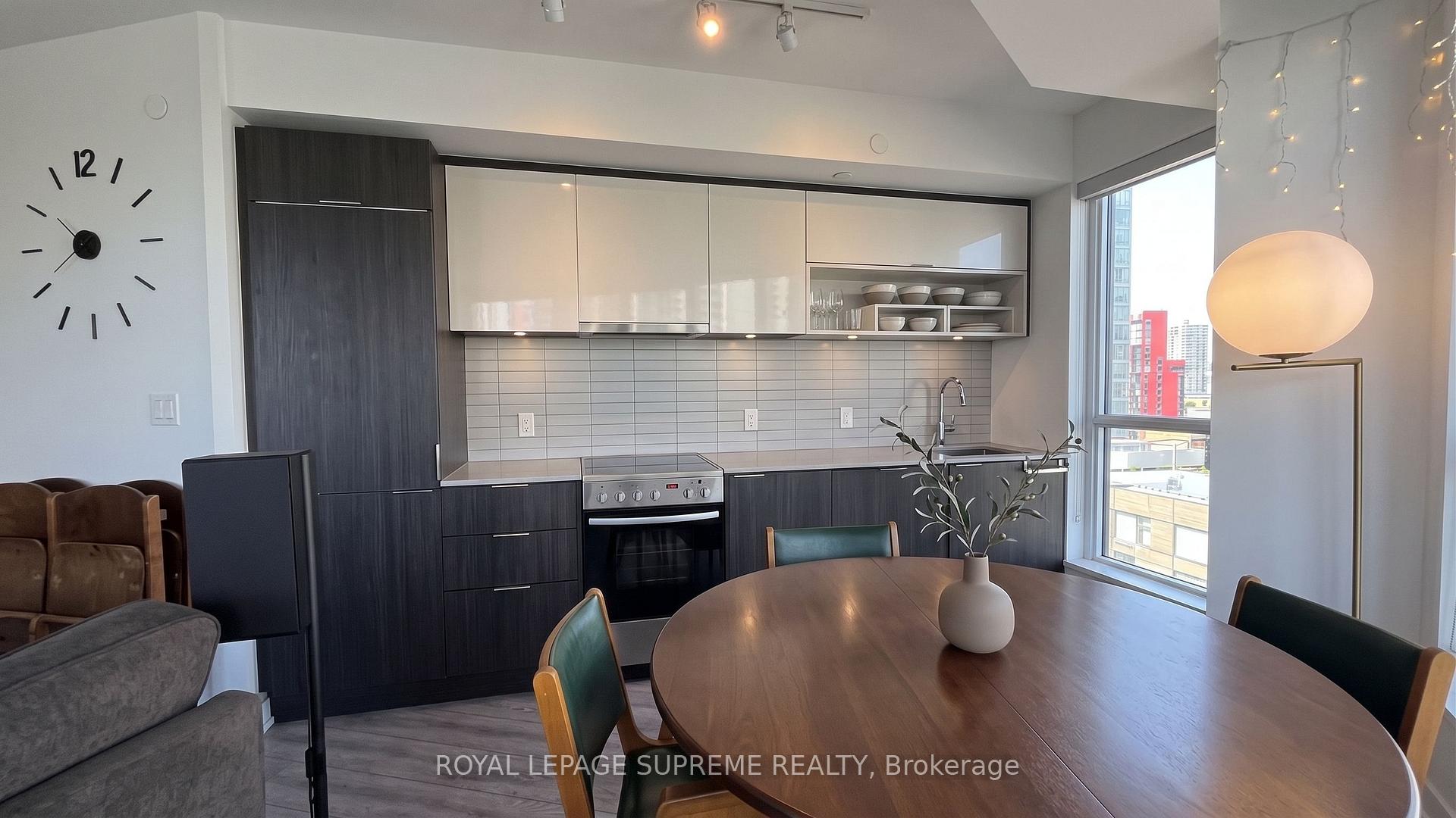 Condo kitchen and dining area with dark cabinetry, a round wood table, and a large window overlooking nearby buildings.