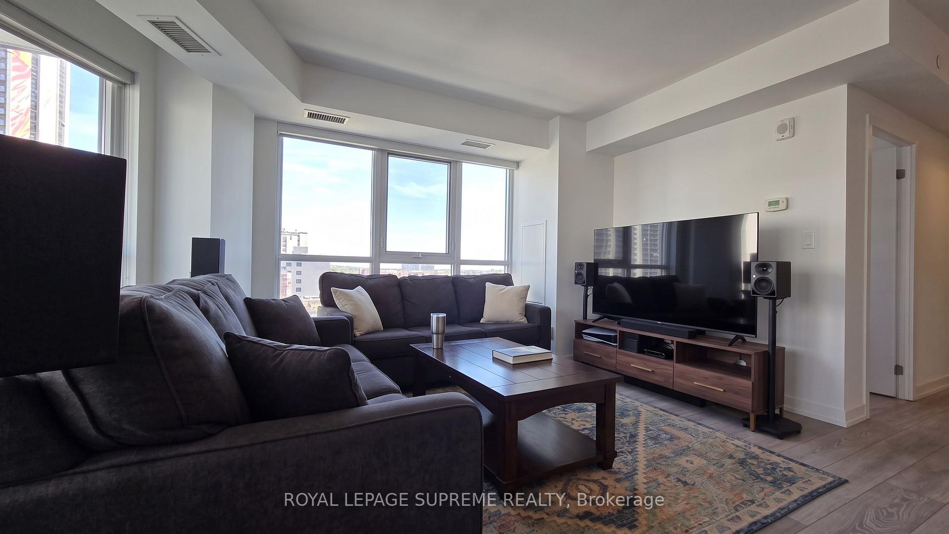 Condominium living room with dark sofas, a large television, and bright windows overlooking nearby buildings.