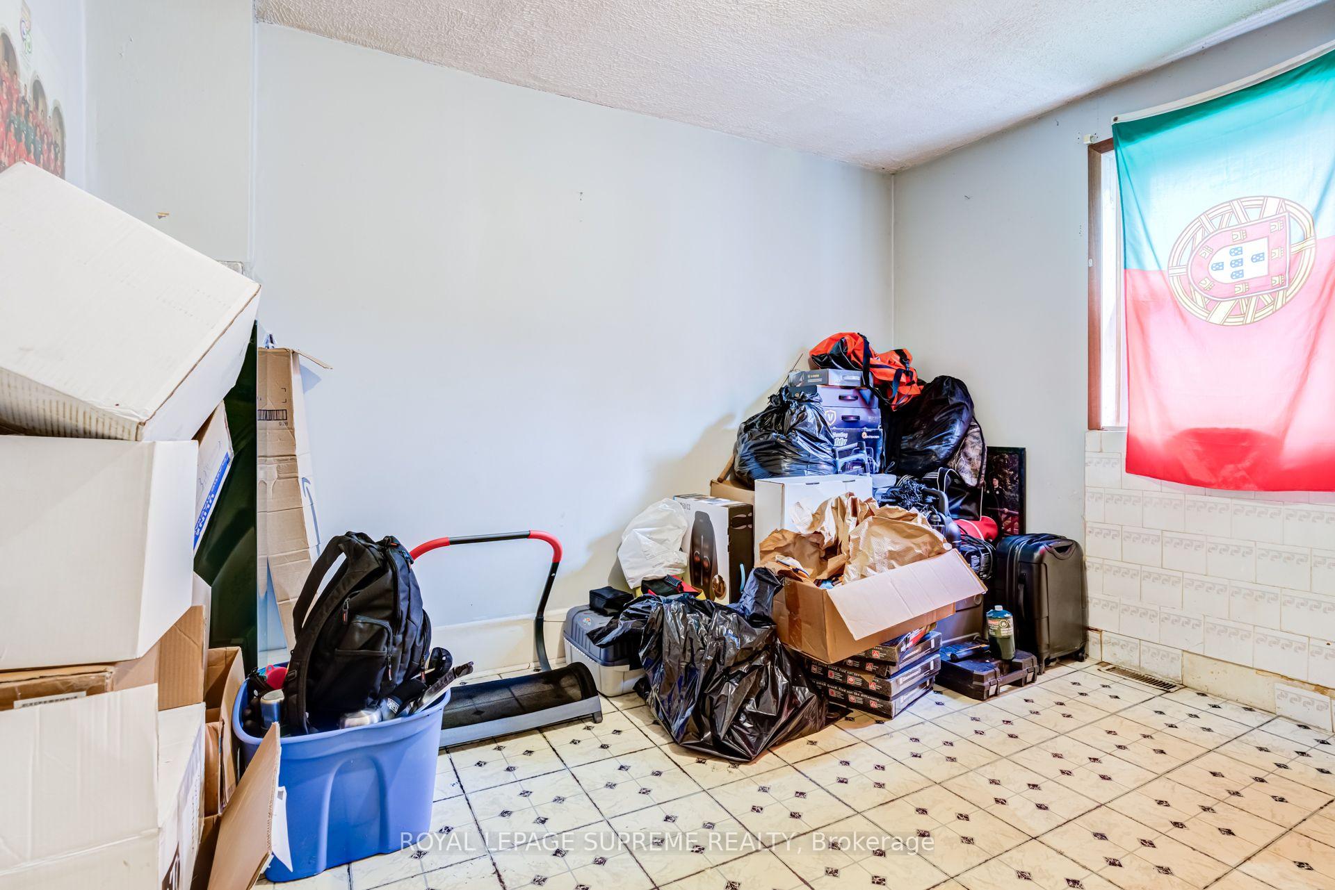 Cluttered room filled with boxes, bags, and luggage beside a window draped with a colorful flag.