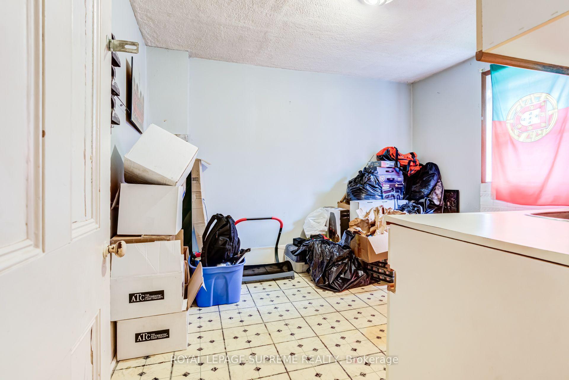 Cluttered kitchen with tiled flooring, white counters, and boxes and bags stacked around the room.