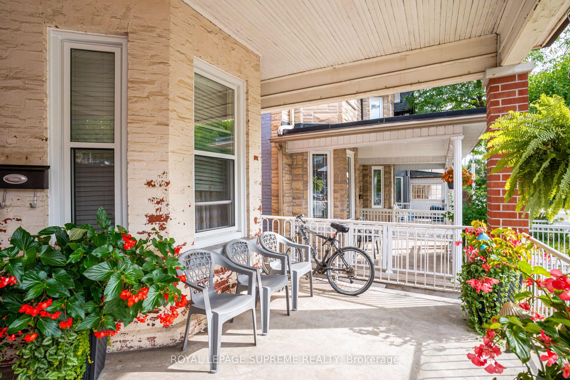 Covered front porch with white railings, outdoor chairs, a bicycle, and abundant flowering plants.