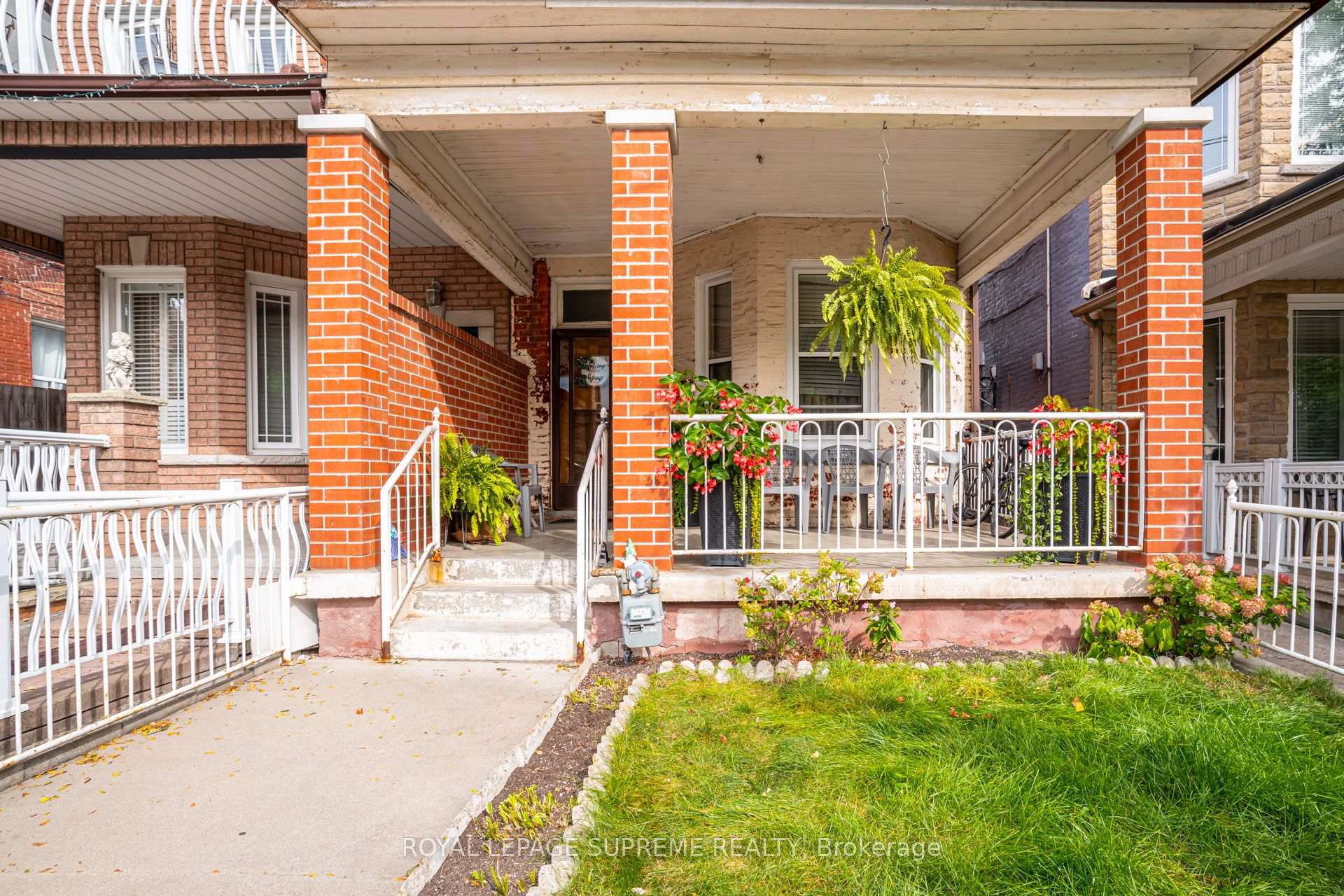 Brick home with a covered porch, white railings, hanging ferns, and flowering plants.