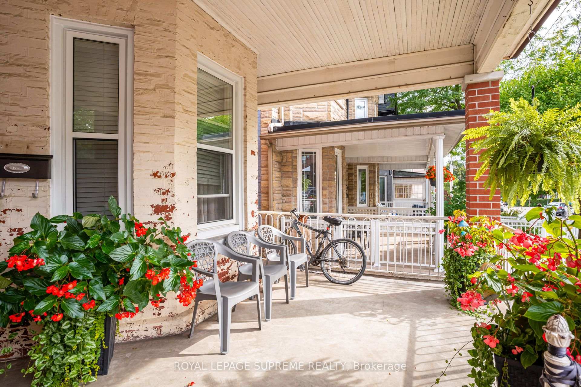 Covered front porch with white railings, patio chairs, a bicycle, and flower-filled planters.