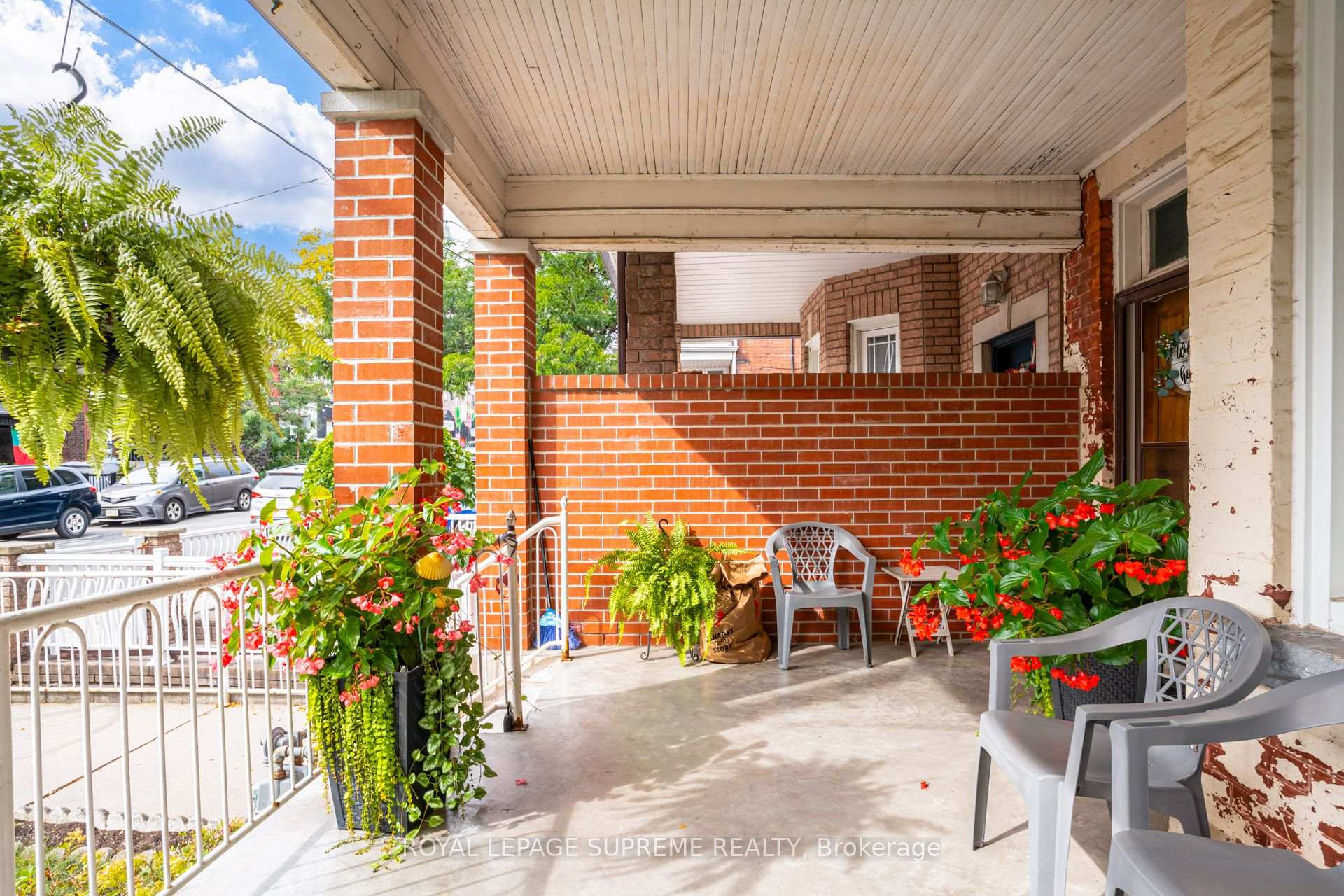 Covered brick front porch with patio chairs, hanging ferns, and red flowering plants.