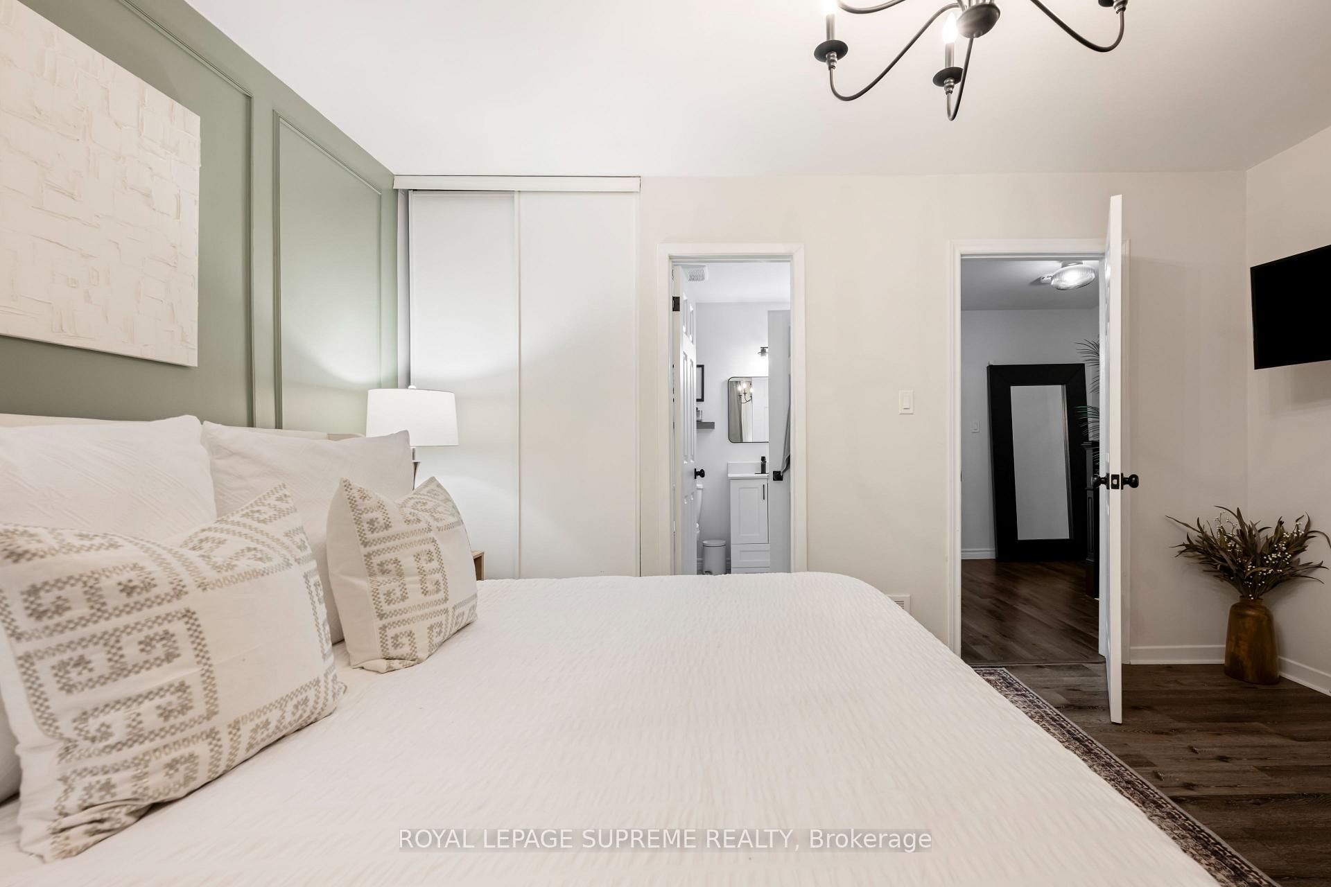 Staged bedroom with a white bed, sage-green paneled wall, and open doors to the bathroom and hallway.