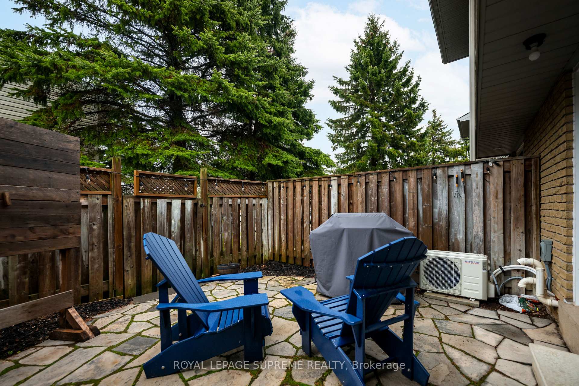 Fenced stone patio with blue Adirondack chairs, a covered barbecue, and tall evergreens.