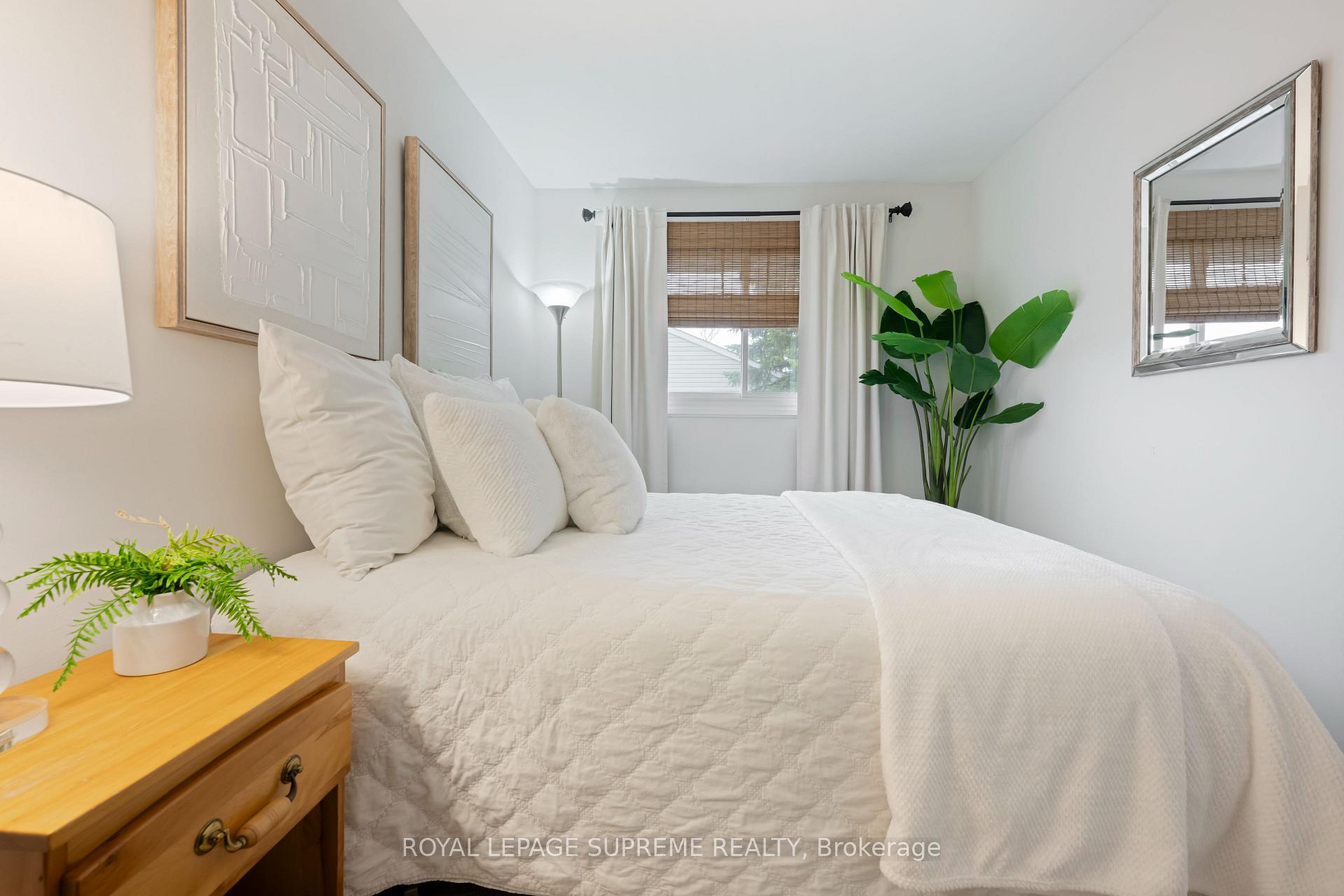 Staged bedroom with a white quilted bed, wooden dresser, woven window blinds, and a tall leafy plant.