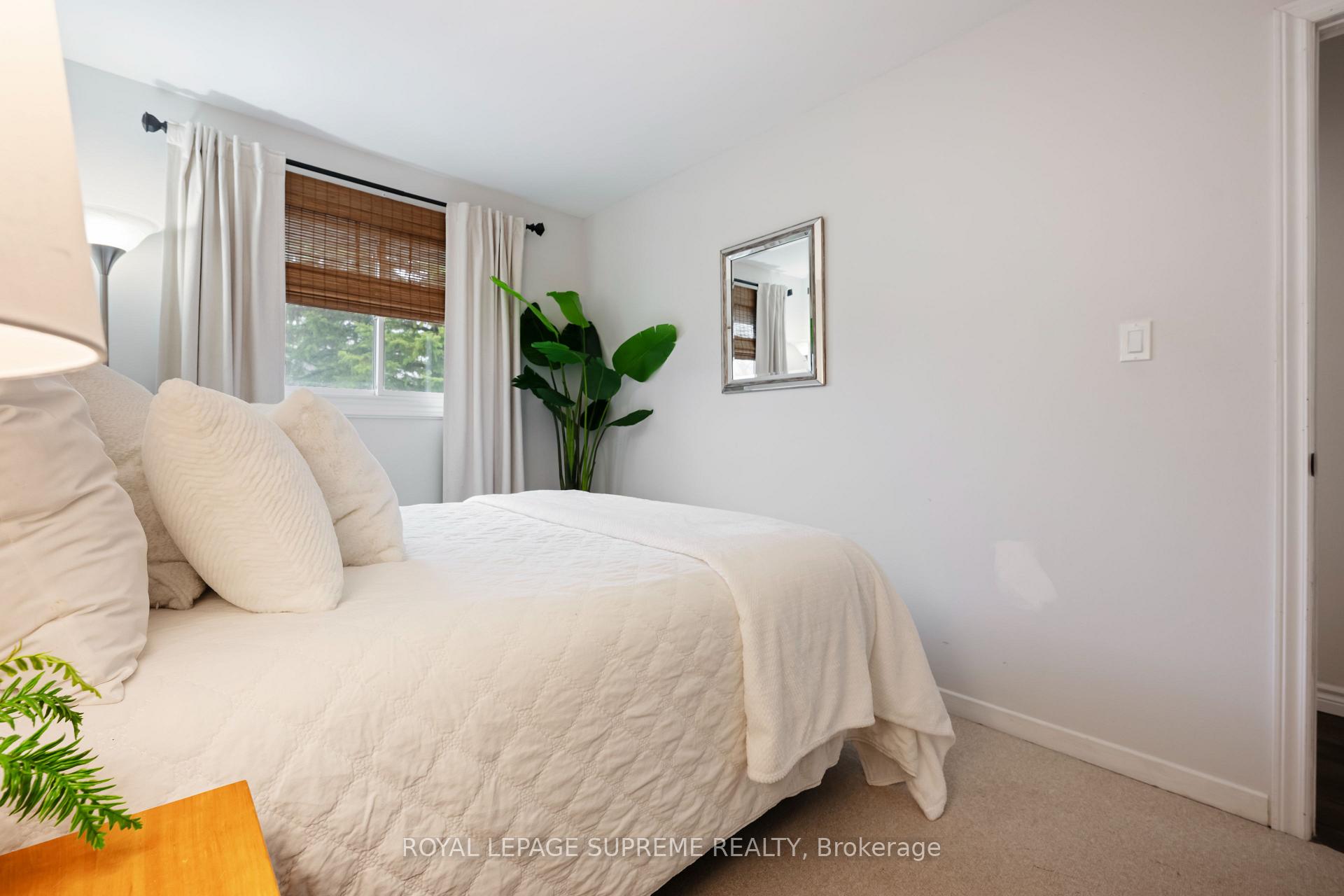 Bedroom with a white bed, bamboo window shade, leafy plant, and framed mirror.