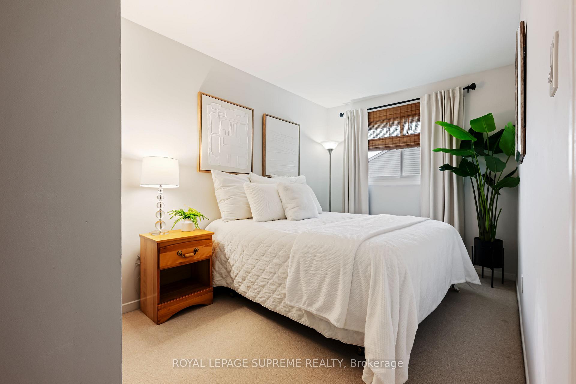 Staged bedroom with a white bed, wooden bedside table, curtained window, and large potted plant.