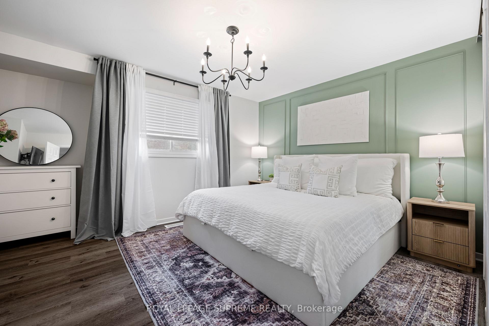 Staged bedroom with a white bed, sage-green paneled wall, chandelier, and patterned rugs.