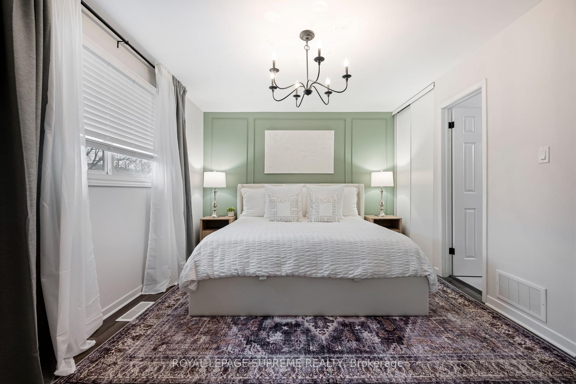 Staged bedroom with a white bed, sage-green paneled wall, chandelier, and patterned rug.
