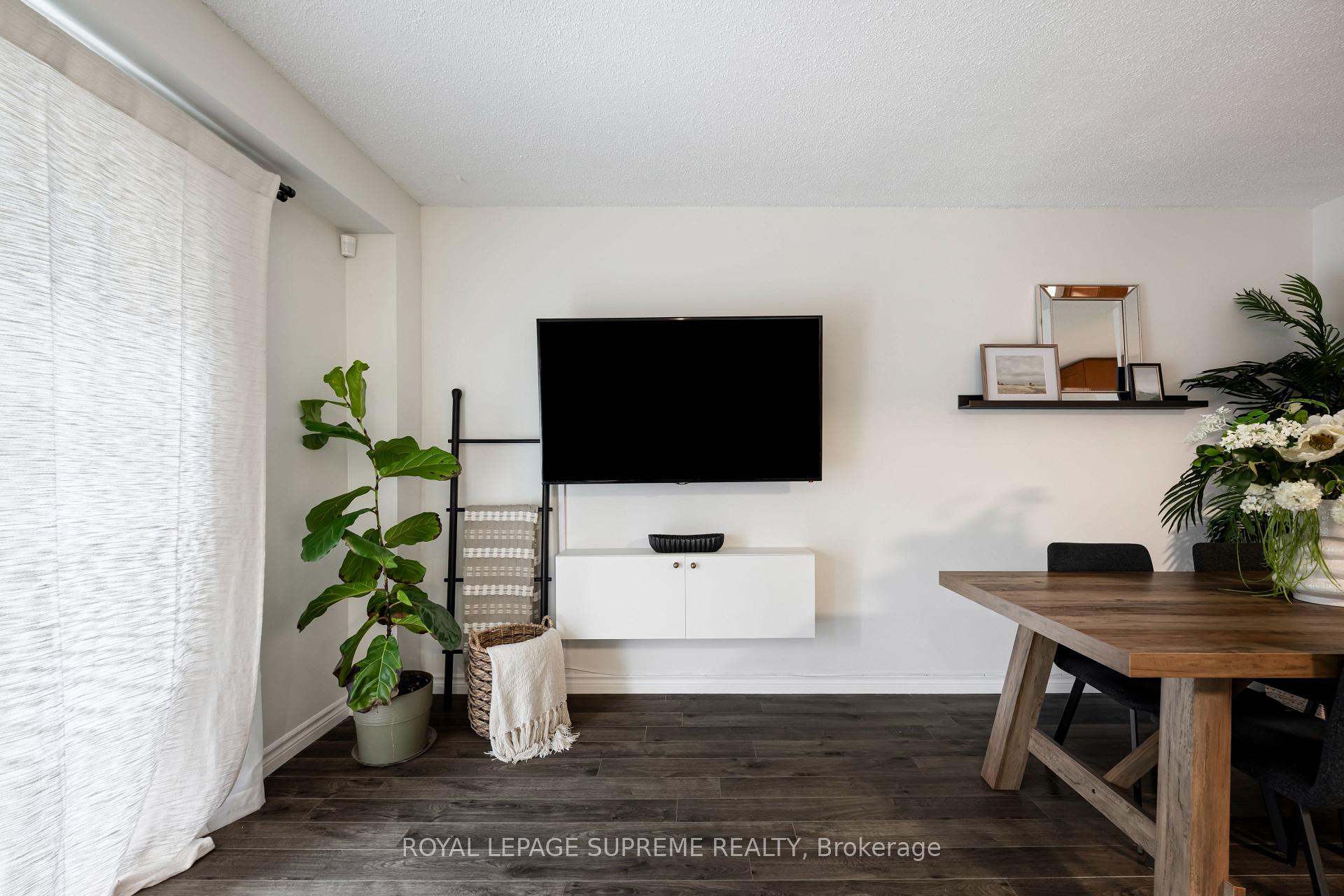 Living and dining area with a wall-mounted TV, white cabinet, large plants, and dark wood flooring.