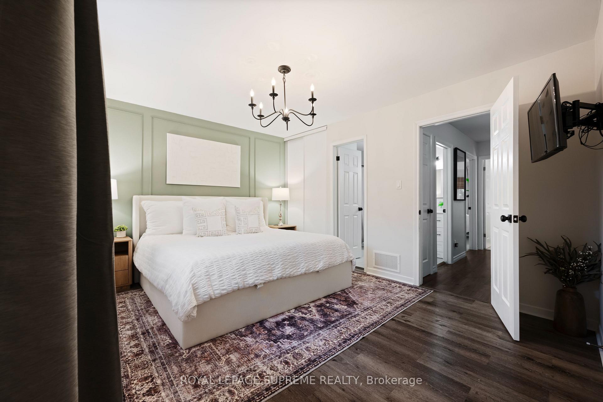 Staged bedroom with a white bed, sage-green paneled wall, patterned rug, and dark wood flooring.