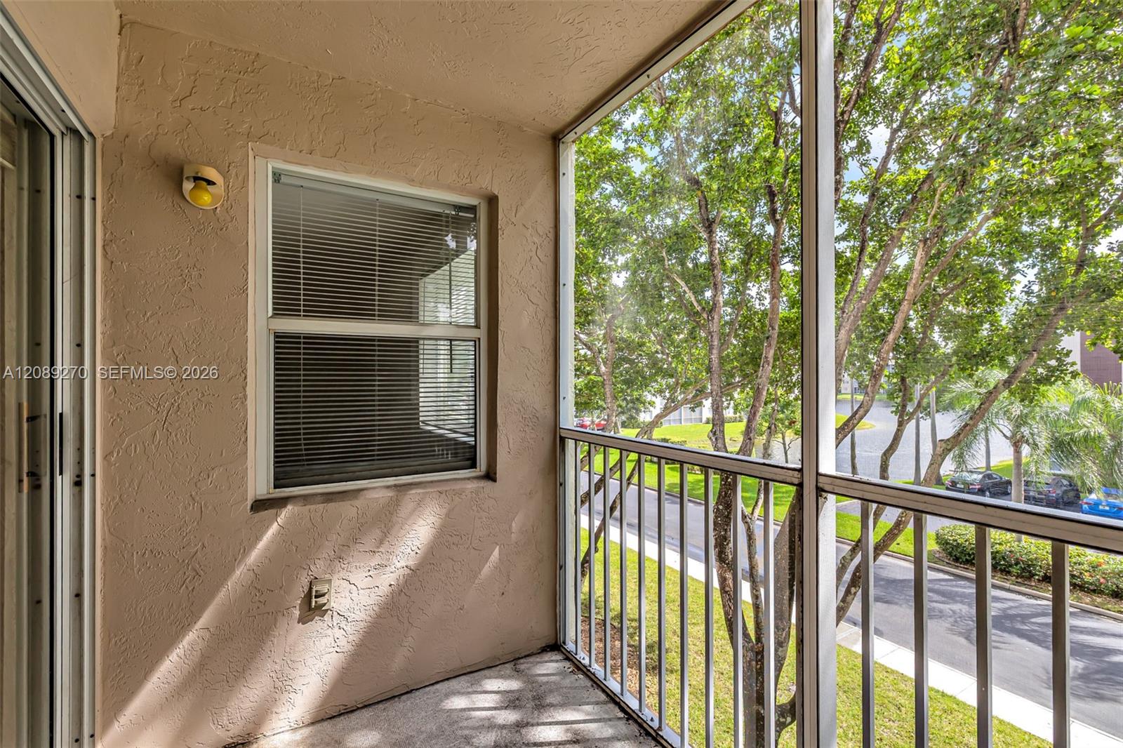 Screened balcony with a stucco wall and leafy trees visible beyond the railing.