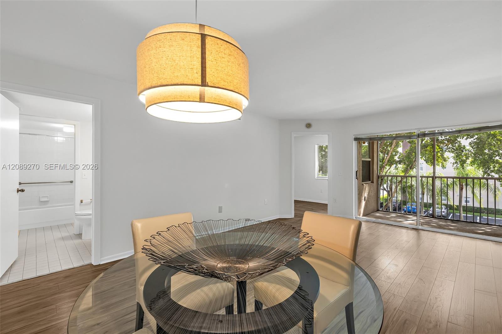 Condominium dining area with a glass table, tan chairs, and a balcony overlooking leafy trees.