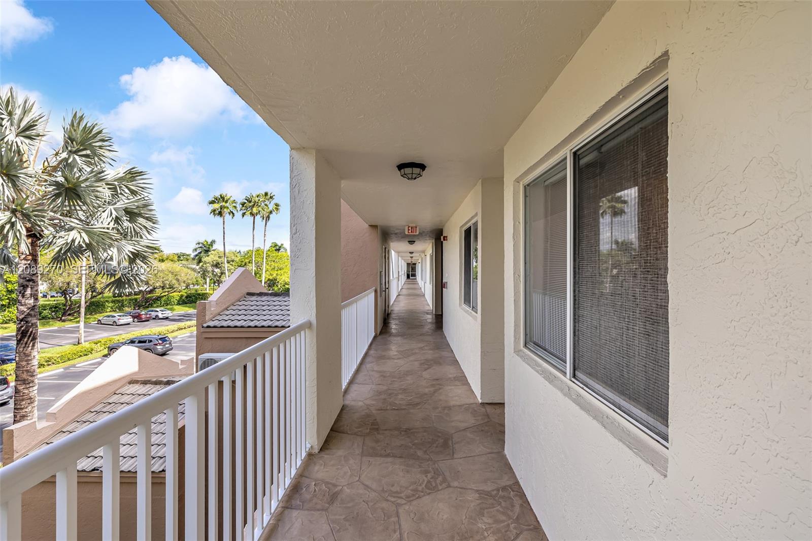 Covered tiled balcony corridor with a white railing overlooking palm trees and a parking lot.