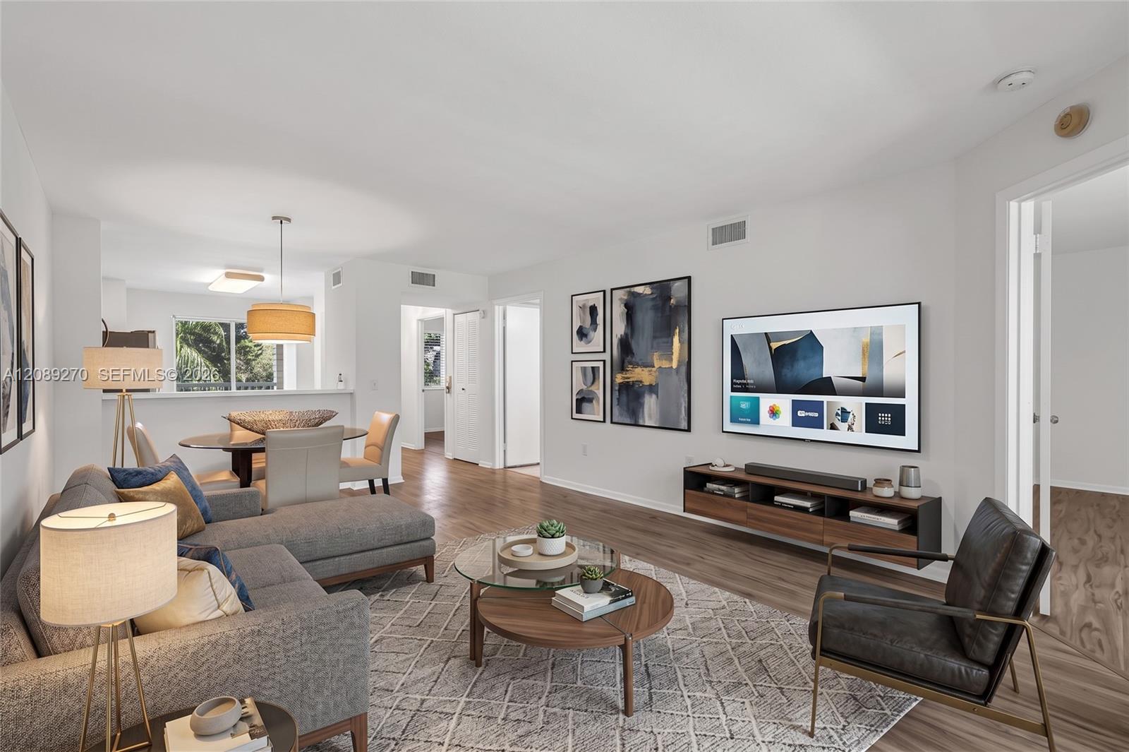 Furnished condo living room with a gray sectional, patterned rug, glass coffee table, and open dining area.