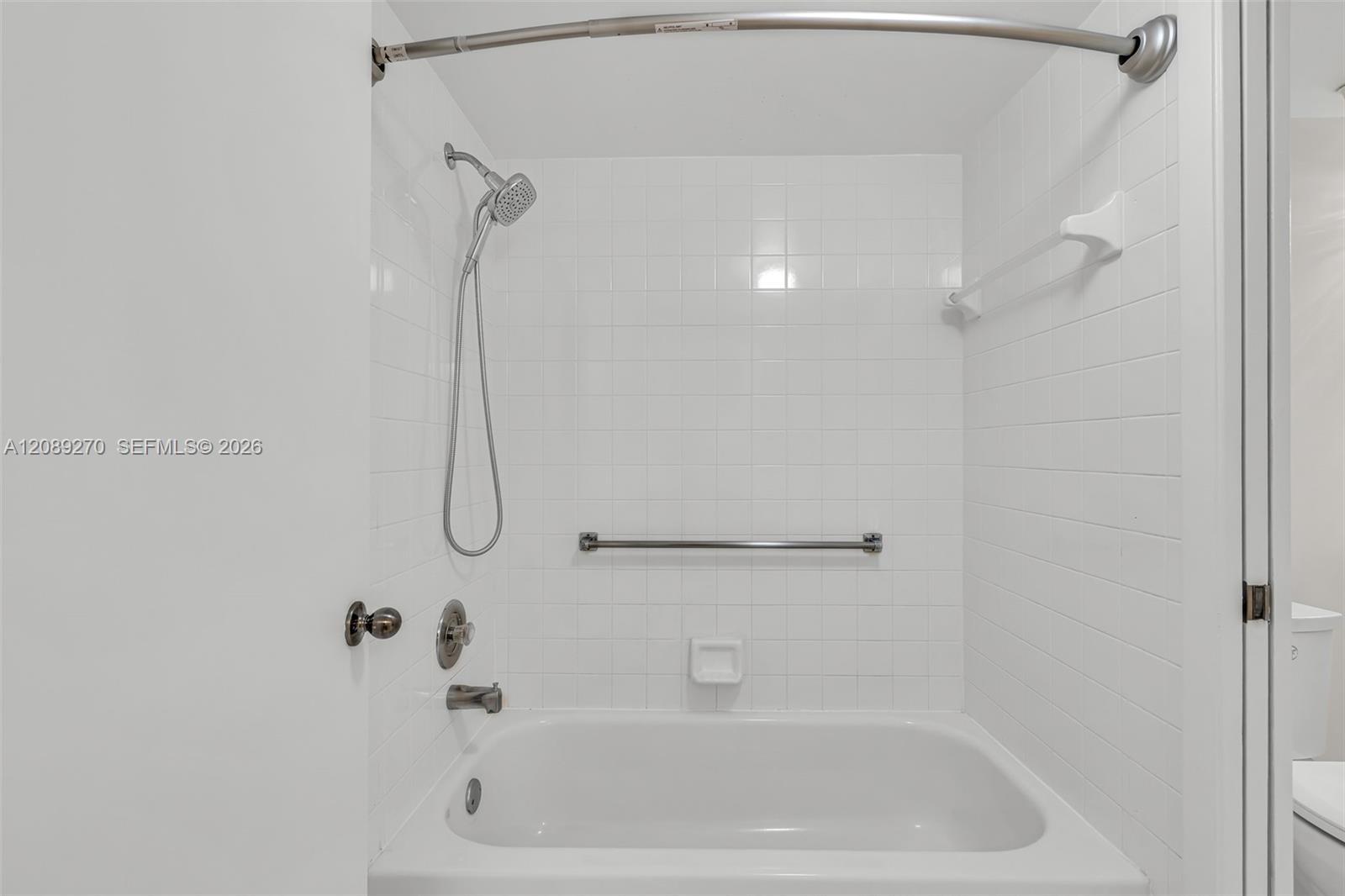White-tiled bathroom with a bathtub, handheld showerhead, and grab bars.