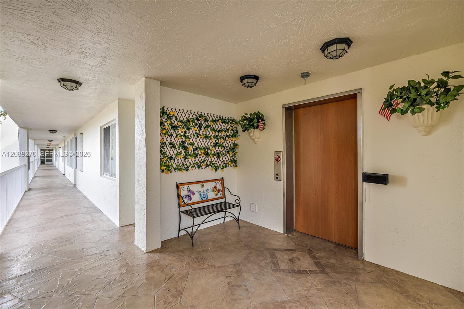 Covered condominium hallway with a wooden elevator door, patterned tile floors, and a decorative bench.