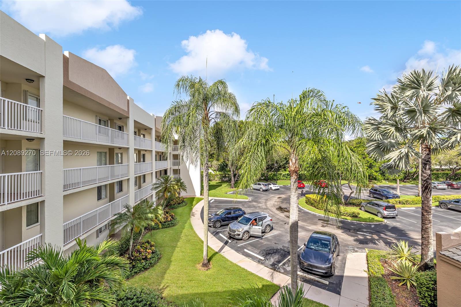 Condominium building with white balconies, palm trees, and a landscaped parking lot.