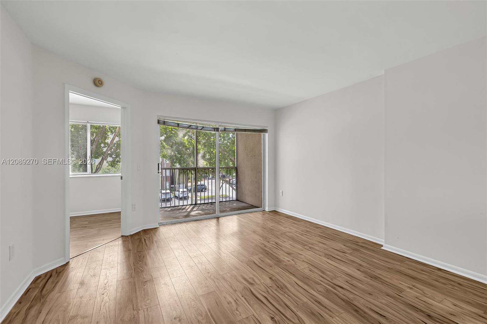 Empty living room with wood-look floors, a sliding glass door to a balcony, and an adjoining room.