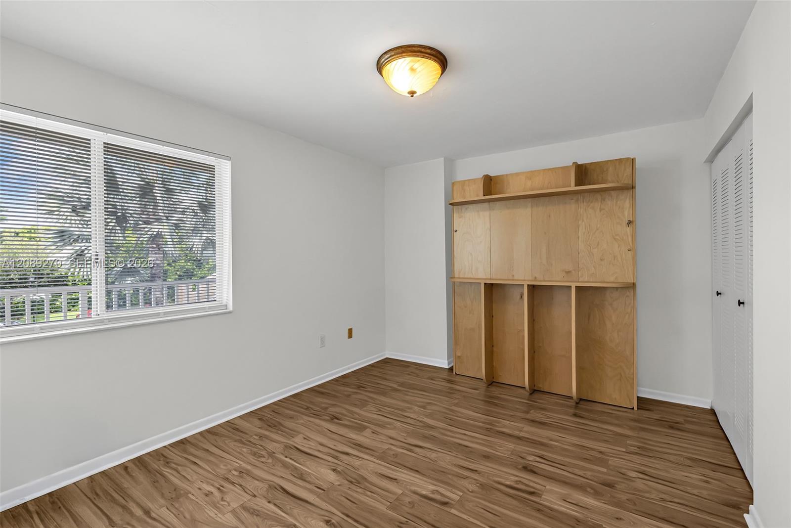 Unfurnished bedroom with wood-look flooring, built-in wooden shelving, and a window overlooking palm trees.