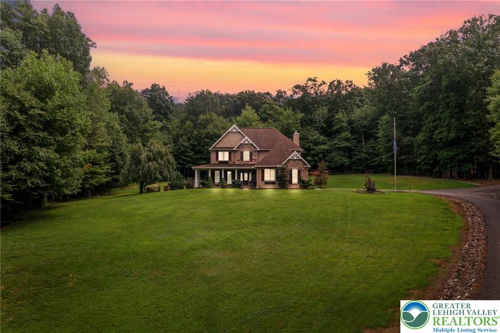 Two-story home amid trees, set behind a broad lawn beneath a pink sunset sky.
