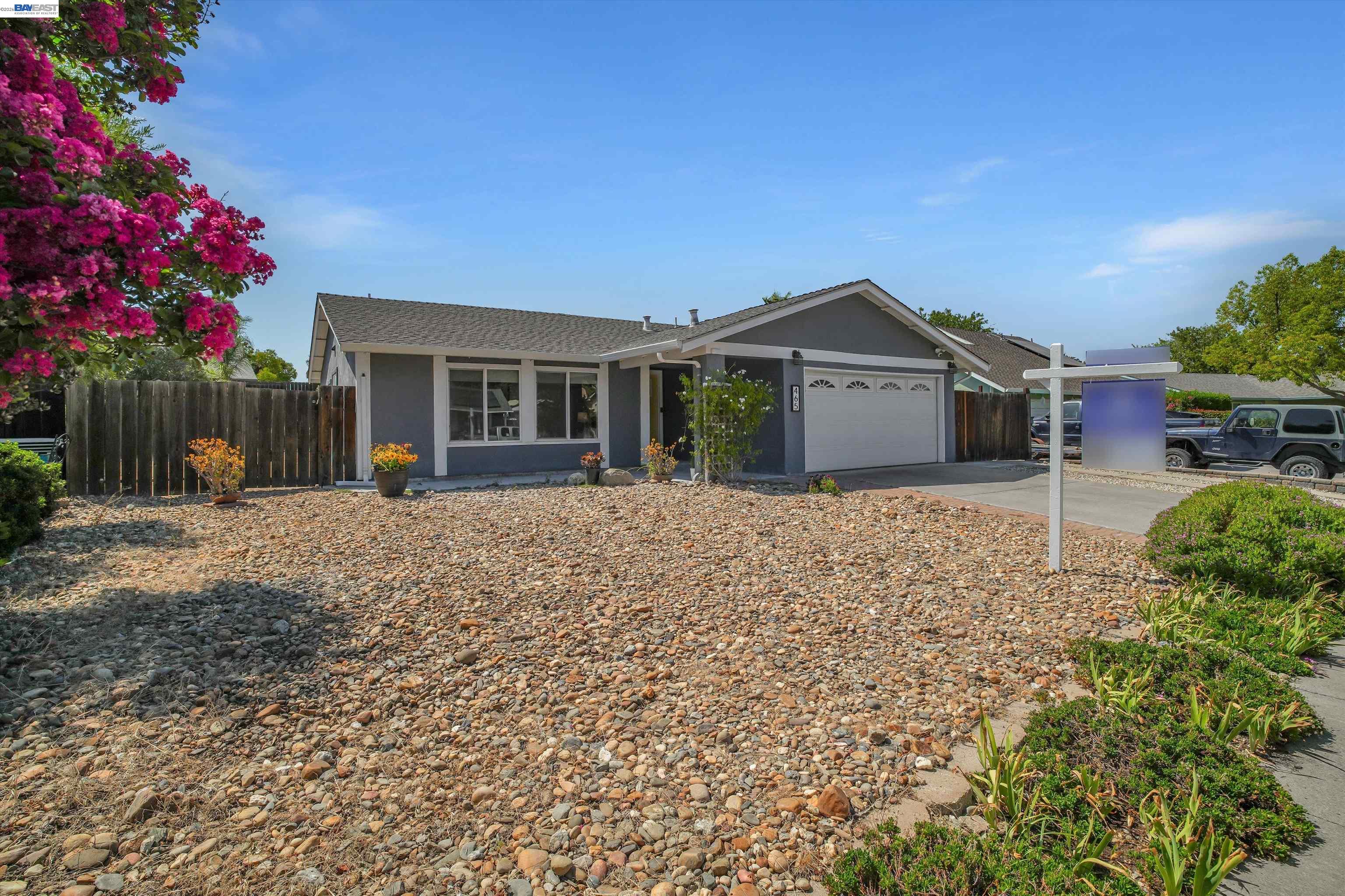 Gray single-story home with an attached garage and rock-covered front yard beneath flowering trees.