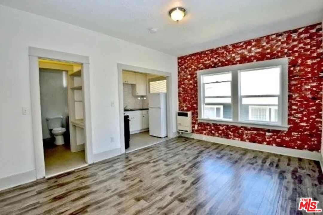 Vacant living room with red patterned accent wall, wood-look flooring, and kitchen and bathroom doorways.