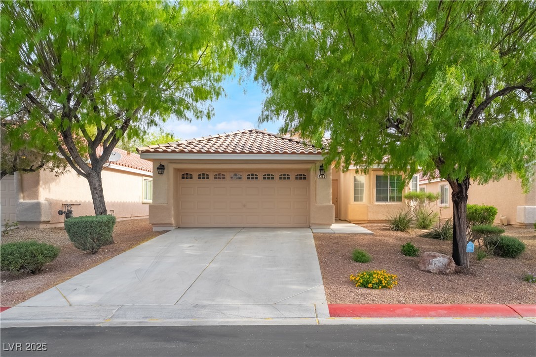 Stucco home with a tiled roof, two-car garage, and desert landscaping beneath mature trees.