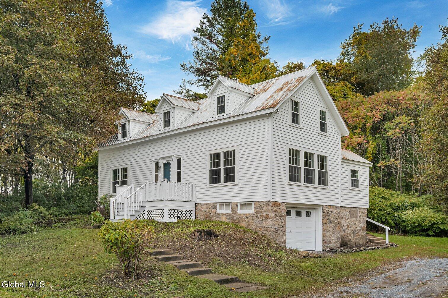 White clapboard house with a stone foundation, metal roof, and raised porch among autumn trees.