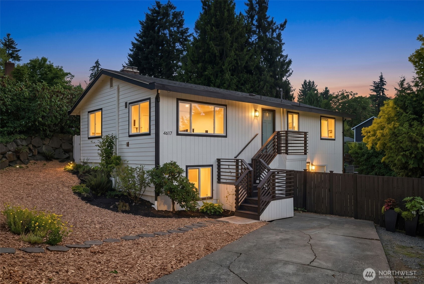 White two-level house at dusk with glowing windows, dark exterior stairs, and landscaped grounds.