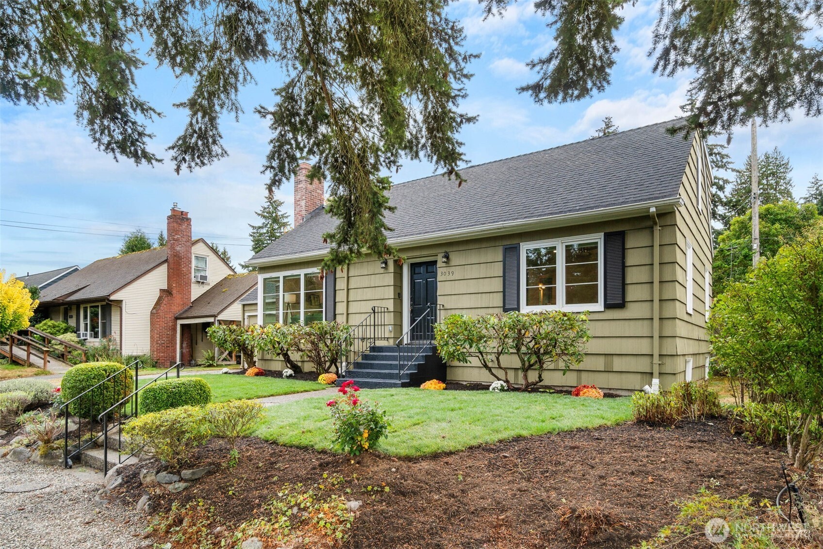 Green shingle-sided house with a steep roof, brick chimney, and landscaped front lawn.