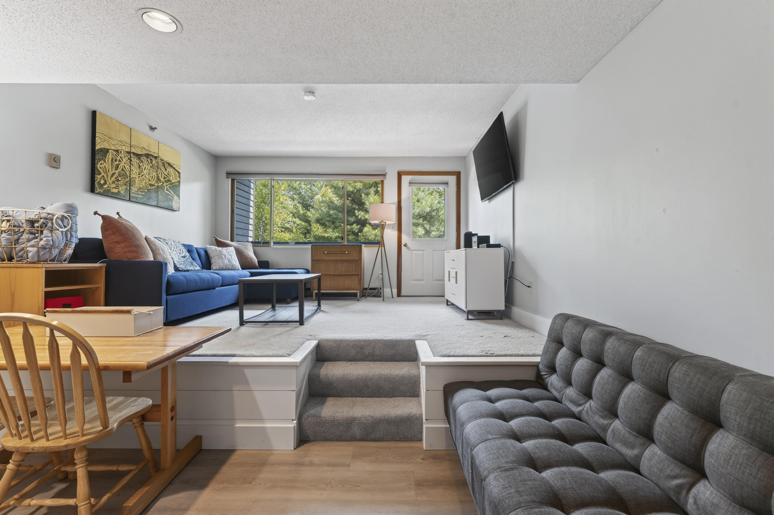 Furnished den with blue and gray sofas, a wall-mounted TV, and carpeted steps to the raised seating area.