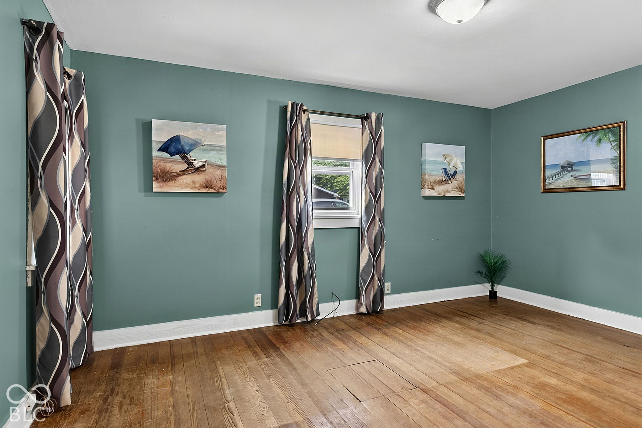 Empty room with teal walls, hardwood floors, patterned curtains, and beach-themed wall art.