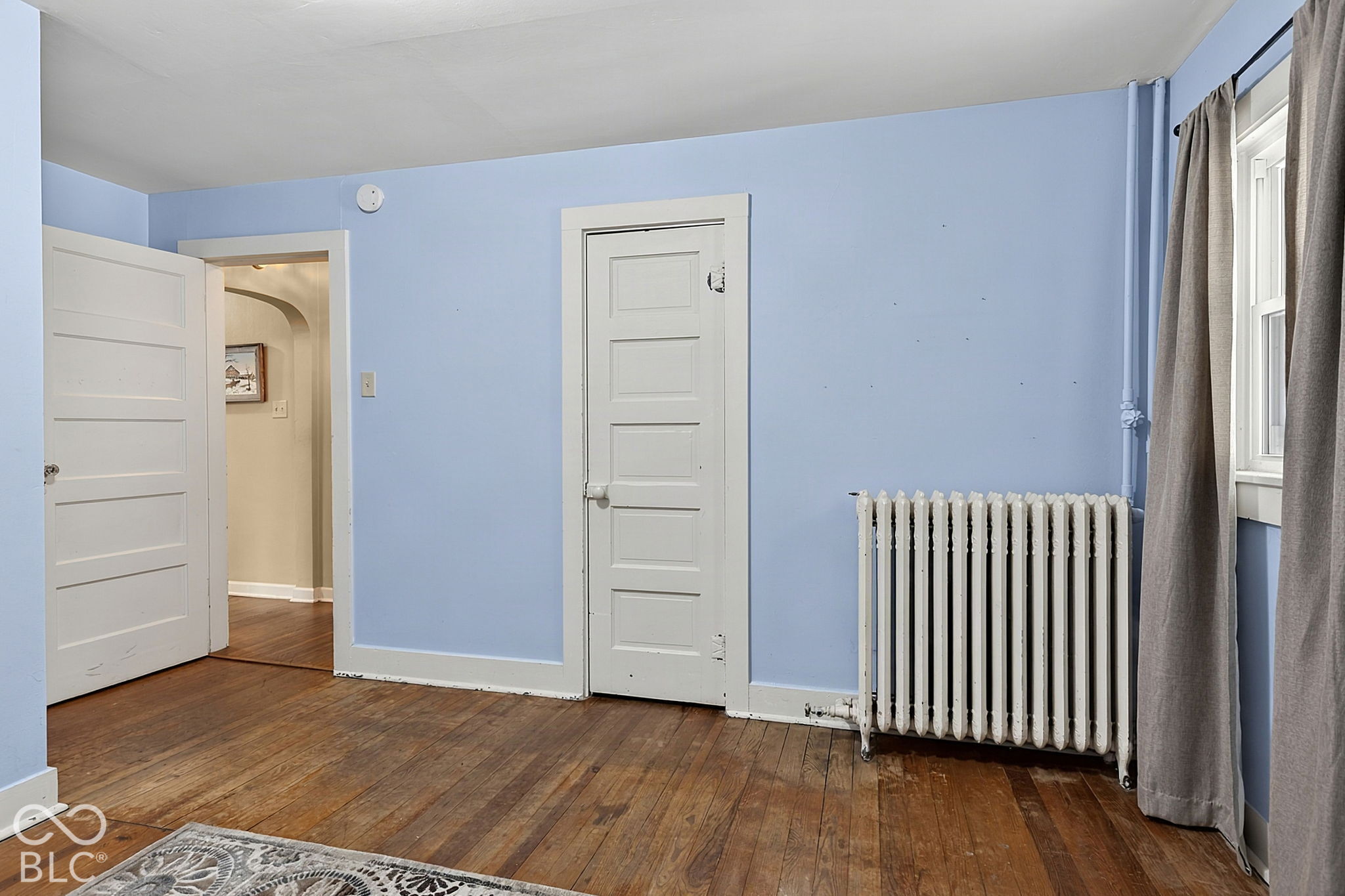 Empty blue-walled room with hardwood floors, a white radiator, and a curtained window.
