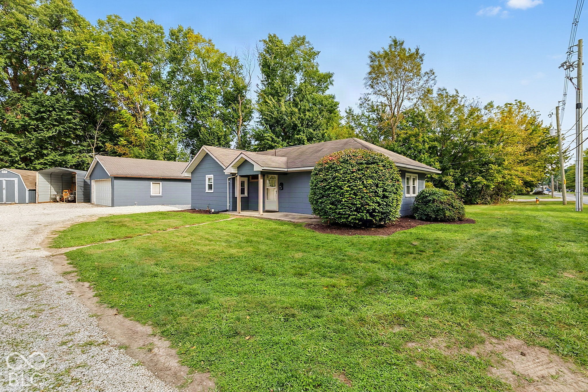 Blue single-story house with a covered entry, garage wing, sheds, and gravel driveway beside a broad lawn.
